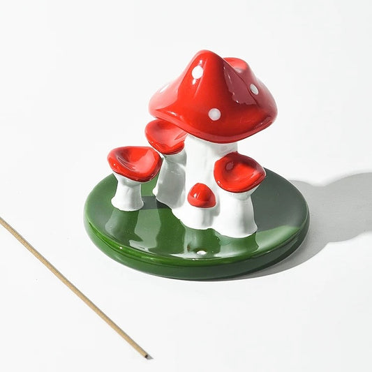 Unlit red and white ceramic mushroom backflow incense burner on a green leaf base with a wooden incense stick lying beside it