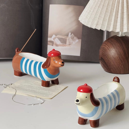 Two ceramic dachshund incense holders in blue striped outfits with red berets on a desk; the brown one holds a lit incense stick, lamp and framed art in the background.