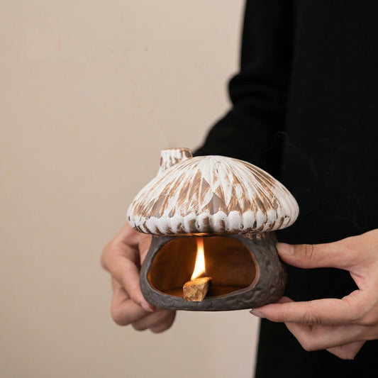 Hands holding a rustic ceramic mushroom house incense burner with a lit palo santo stick burning inside showing a warm flame