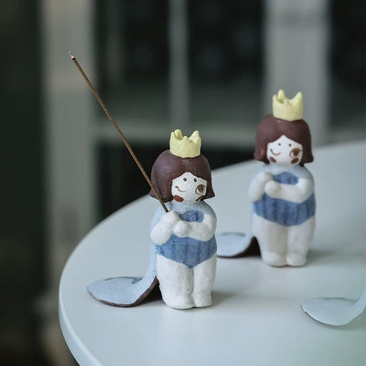 Two handmade ceramic girl incense holders with yellow crowns and blue outfits on a white surface; the front figure holds an inserted incense stick with a leaf-shaped ash tray behind.