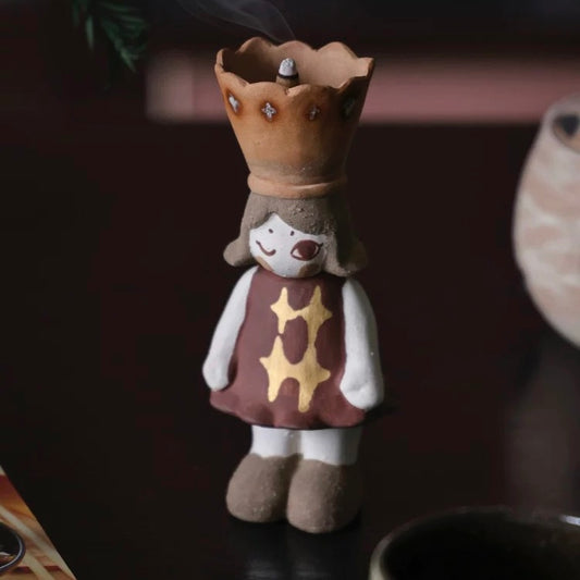 Handcrafted ceramic girl figurine incense burner with a crown-shaped top for cone incense, star-patterned dress, standing on a dark background.