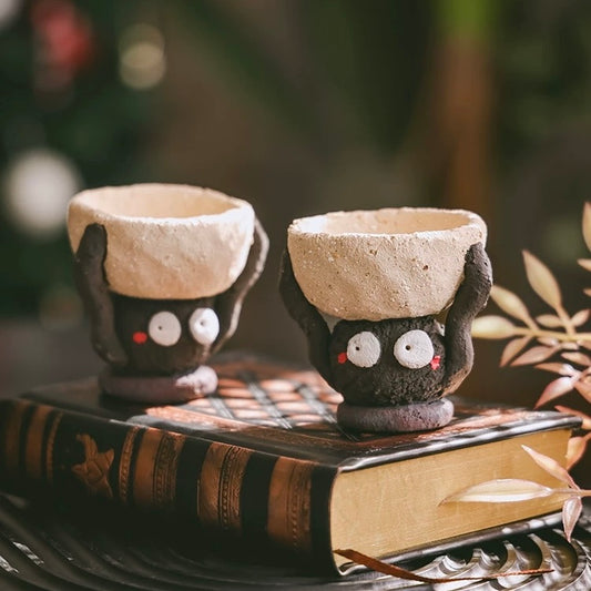 Two handmade ceramic candle holders shaped like small figures holding textured bowls overhead, displayed on vintage books with greenery; designed to hold tealight candles.
