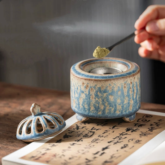 Handmade blue ceramic electric incense burner with the lid off while incense powder is added above a calligraphy scroll.