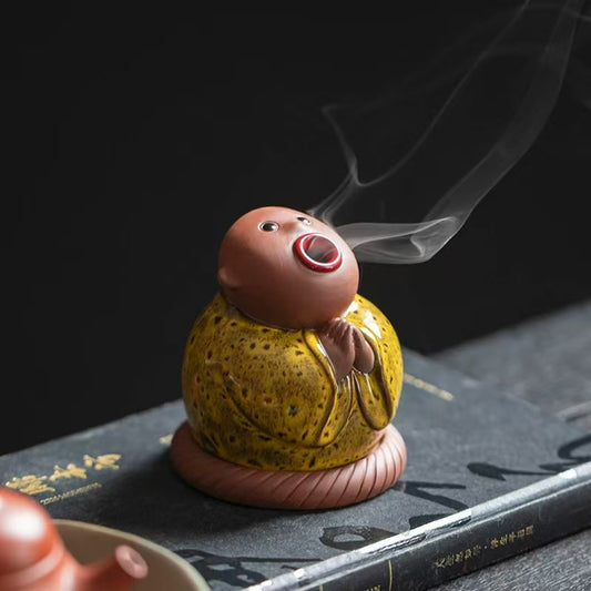 Abstract ceramic incense burner shaped like a big mouth monk with smoke flowing from the mouth, sitting on a black surface