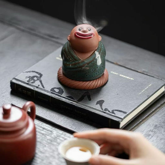 Abstract ceramic incense burner featuring a smiling monk in a green robe with smoke flowing from the mouth, placed on a black book near a teacup