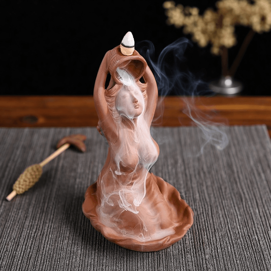 Ceramic goddess-shaped backflow incense burner in use, with smoke cascading down the figure’s body and incense tool beside on fabric mat