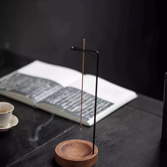 Backflow incense burner with wooden base and hanging stick holder on a dark tea table, open book and teacup in background, smoke rising