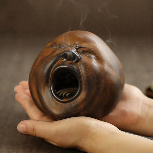 A hand holding a handmade brown-glazed ceramic incense burner shaped like a yawning face, with smoke drifting from the nostrils.