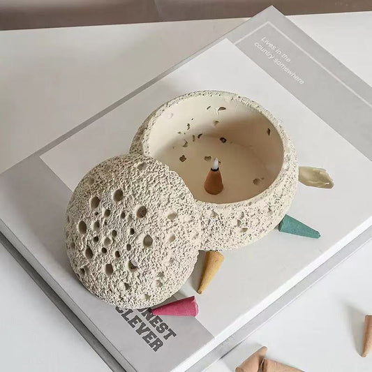 Handmade cement incense burner with removable perforated lid, burning incense cone inside, surrounded by colorful backflow incense cones on a white minimalist setup