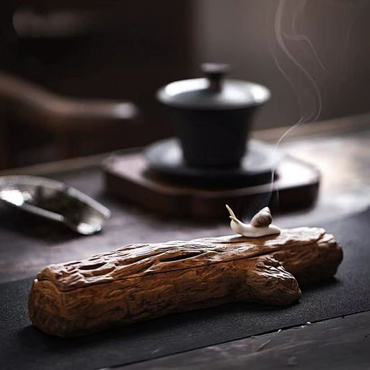 Snail-shaped ceramic incense burner on a rustic wooden incense base, surrounded by a traditional tea set