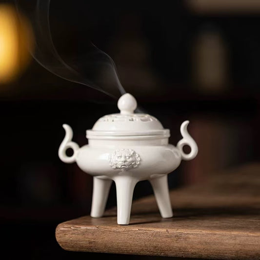 White ceramic tripod incense burner with guardian lion relief and curved smoke, placed on a wooden surface