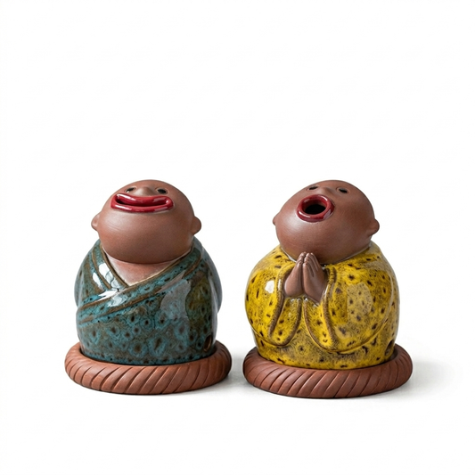 Set of two abstract ceramic monk incense burners with exaggerated facial expressions, featuring one monk in a blue robe and one in a yellow robe, both seated on round bases against a clean white background.