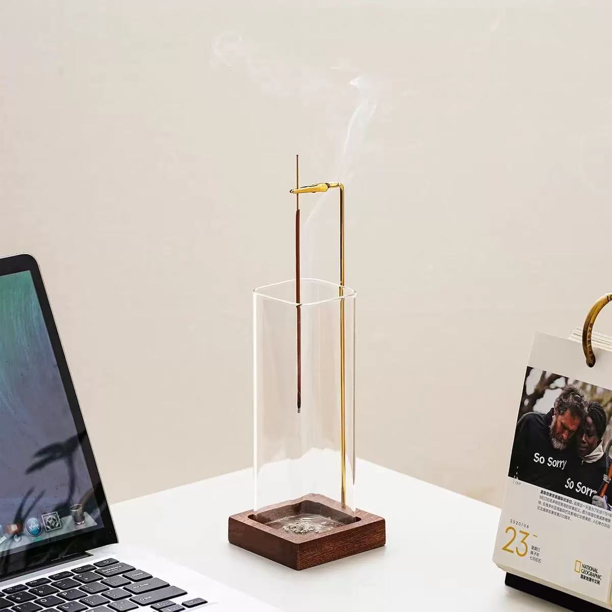 Vertical glass incense holder with wooden base and brass clip placed on a white office desk beside a laptop and calendar
