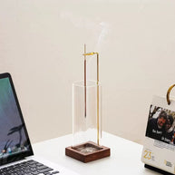 Vertical glass incense holder with wooden base and brass clip placed on a white office desk beside a laptop and calendar
