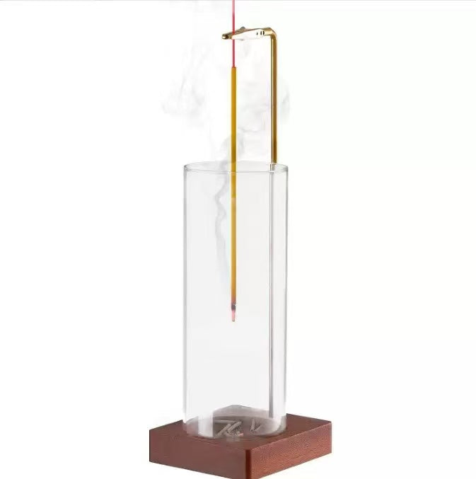 Front view of vertical glass incense holder with a burning incense stick suspended by a brass clip, white background and wooden base visible
