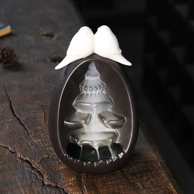 Backflow Incense Burner Ceramic Waterfall with Doves displayed on a rustic wooden surface, featuring two white doves on top and a flowing cascade of backflow incense smoke through the black ceramic waterfall channels