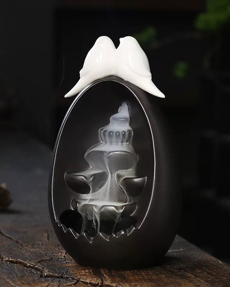 Backflow Incense Burner Ceramic Waterfall with Doves styled on a rustic wooden surface, featuring two white doves on top and a flowing incense smoke waterfall against a moody background with soft blue lights