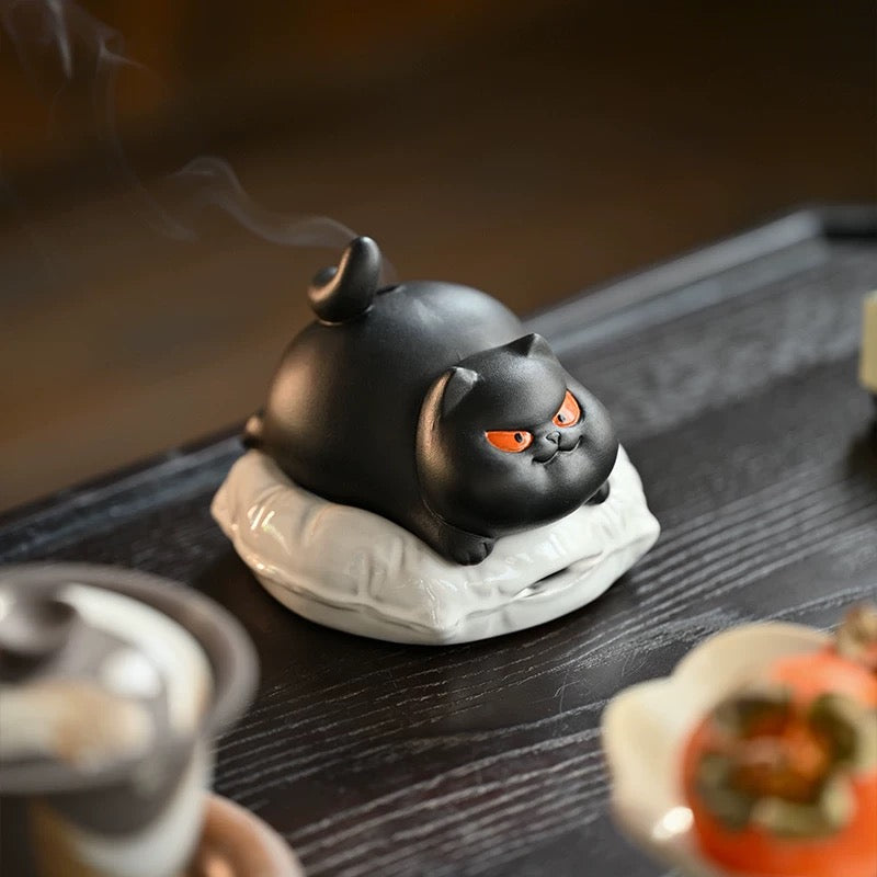 Black ceramic cat incense burner with orange eyes on a white cushion, thin smoke rising, three-quarter angle on a dark wood table with a blurred teacup in the foreground.