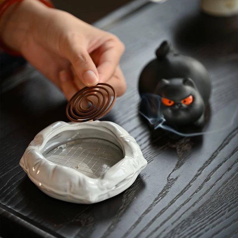 Hand placing a spiral incense coil onto the mesh tray of the white cushion base; black cat ceramic burner in the background with faint smoke.