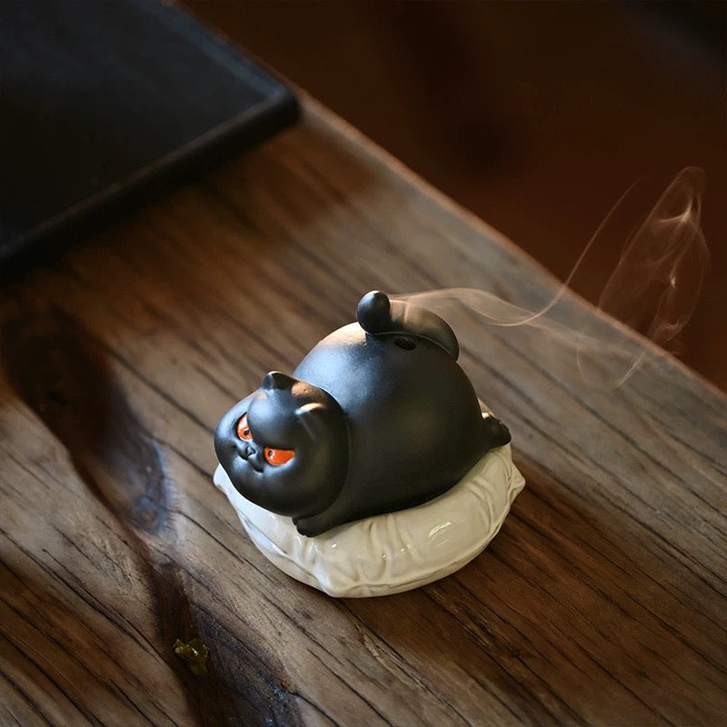 Black ceramic cat incense burner on a white cushion with thin smoke rising on a wooden table.