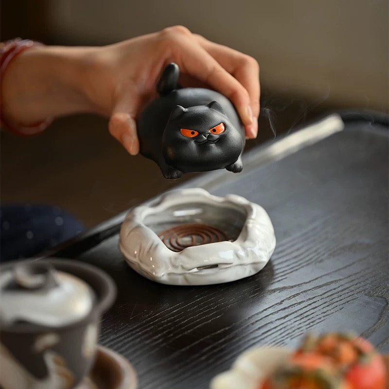 Hand lifting a black ceramic cat incense burner to reveal the white cushion base with a spiral incense coil on a dark wooden table.