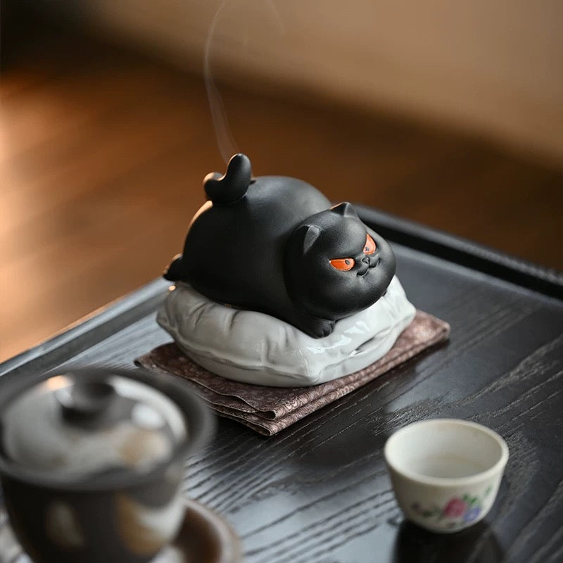 Black ceramic cat incense burner on a white cushion set on a tea tray, thin smoke rising with a small teacup in the foreground.