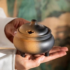 Hand holding a brown and black reactive glaze ceramic electric incense heater with a compact three-leg design for flameless incense use.