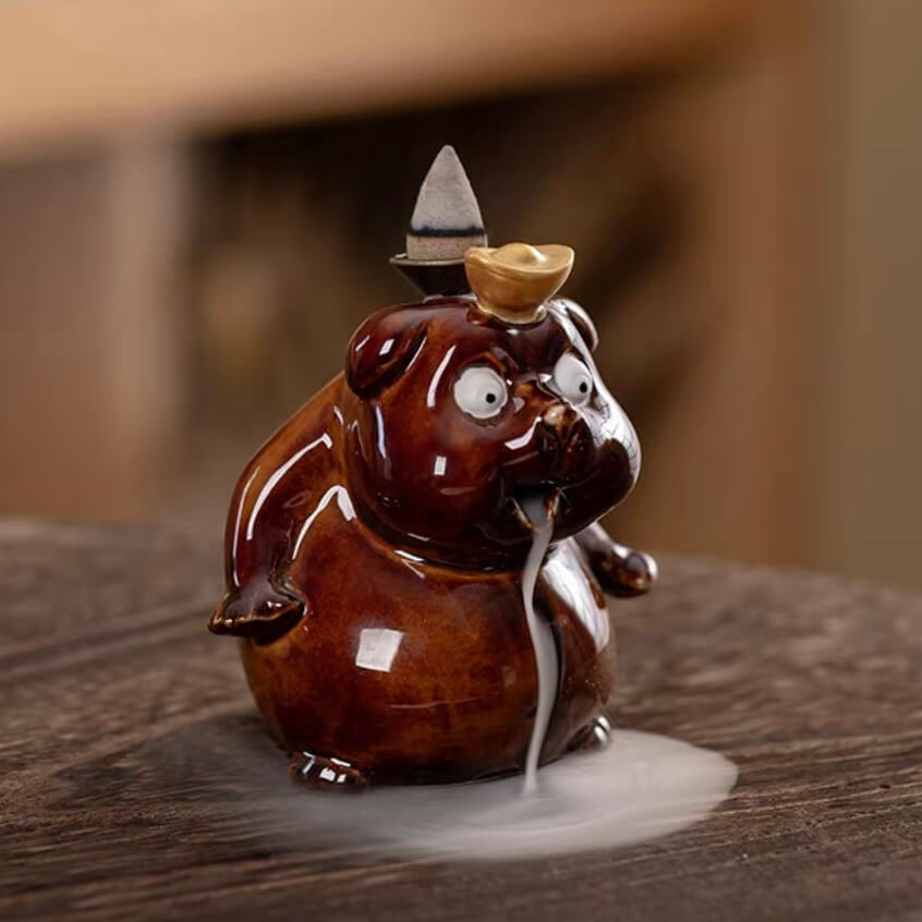 A close-up of a glossy brown ceramic cute animal backflow incense burner sitting on a textured wooden surface, featuring a lit incense cone on its back with a thick trail of white smoke flowing out of its mouth and pooling around its base.