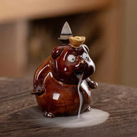 A close-up of a glossy brown ceramic cute animal backflow incense burner sitting on a textured wooden surface, featuring a lit incense cone on its back with a thick trail of white smoke flowing out of its mouth and pooling around its base.