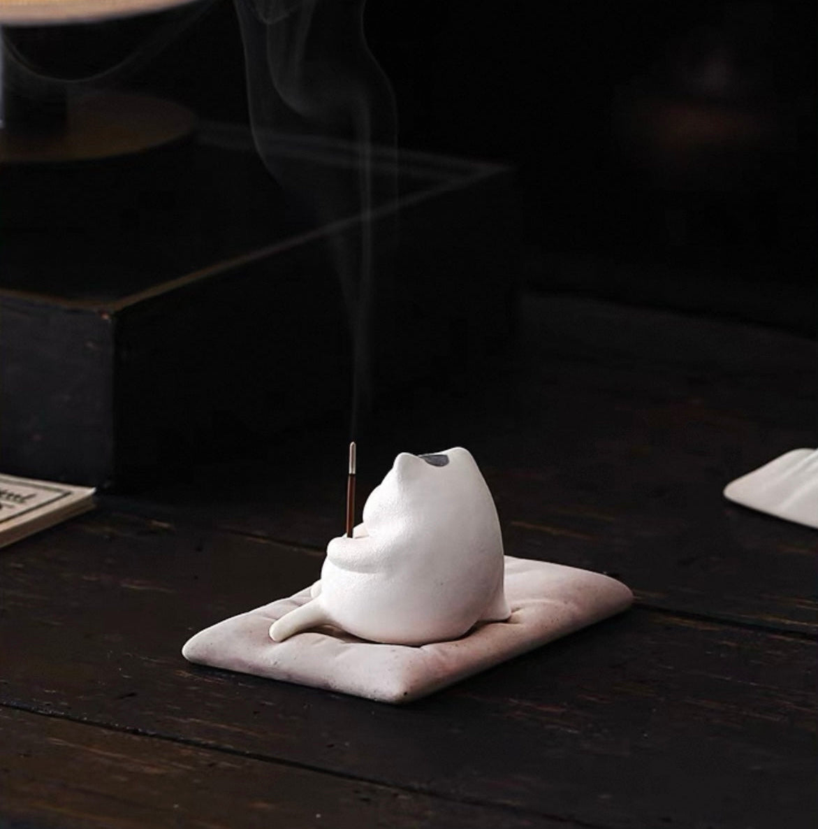 Rear view of a white ceramic sleeping cat incense holder on a cushion with an inserted incense stick; Japanese zen desk decor.