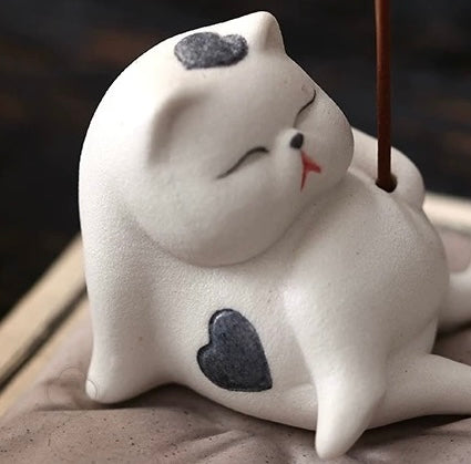 Close-up of a white ceramic cat incense holder on a cushion, heart-shaped markings visible, incense stick inserted.