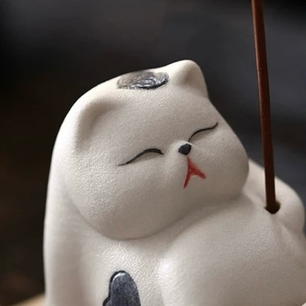 Close-up of the white ceramic cat incense holder’s face with an inserted incense stick on the right; matte texture and heart marking visible.