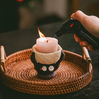 Hand lighting a pillar candle in a handmade ceramic figure candle holder set on a round woven tray.