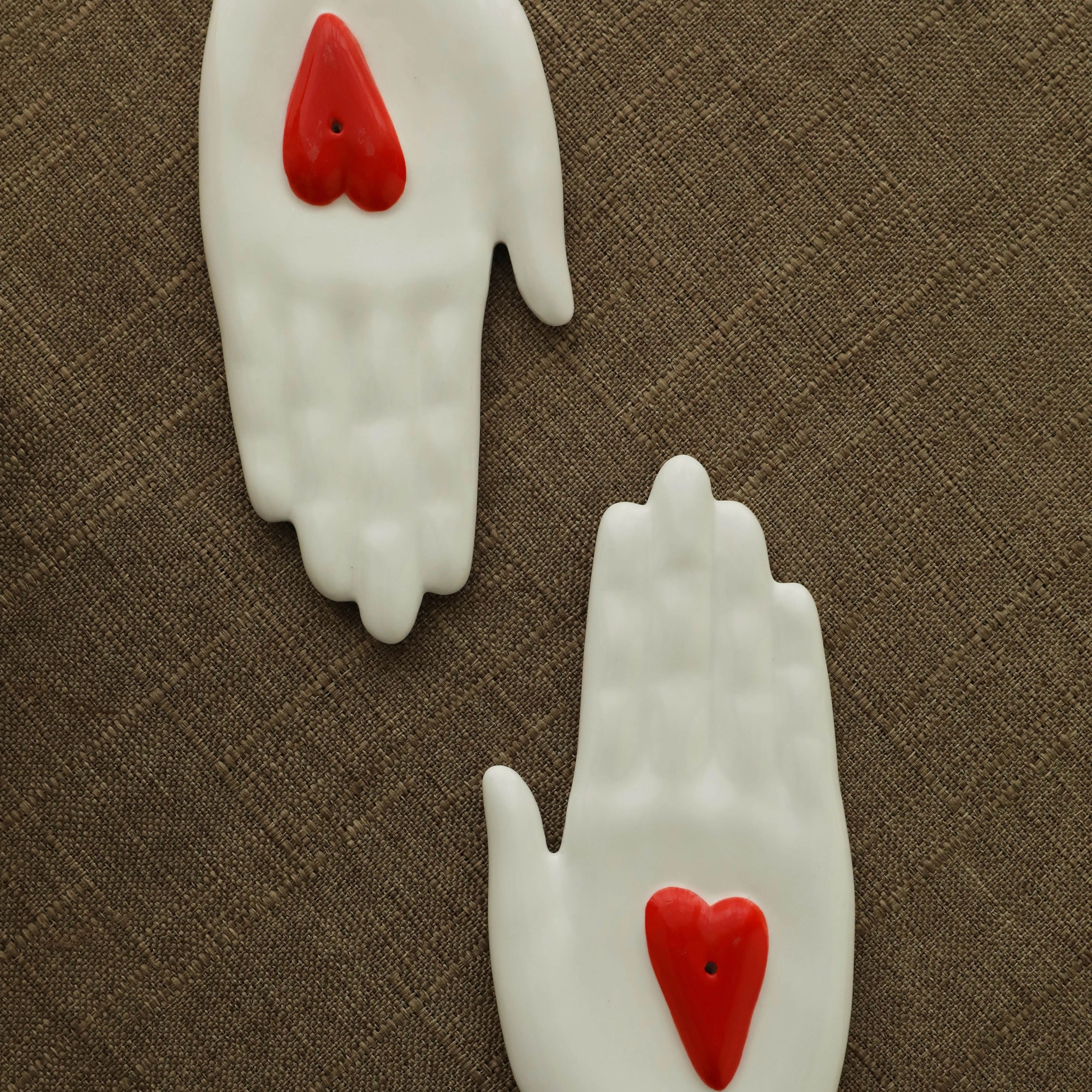 Overhead view of two white ceramic hand-shaped incense holders with red heart inserts on a linen surface.