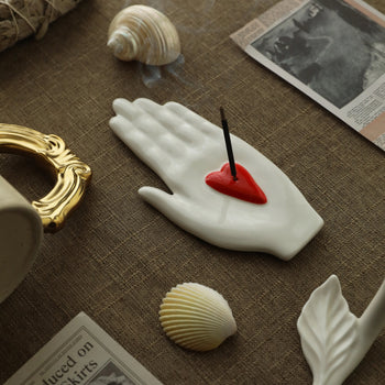 White ceramic hand-shaped incense holder with a red heart insert holding an incense stick, styled with seashells and a small gold frame on a linen surface.