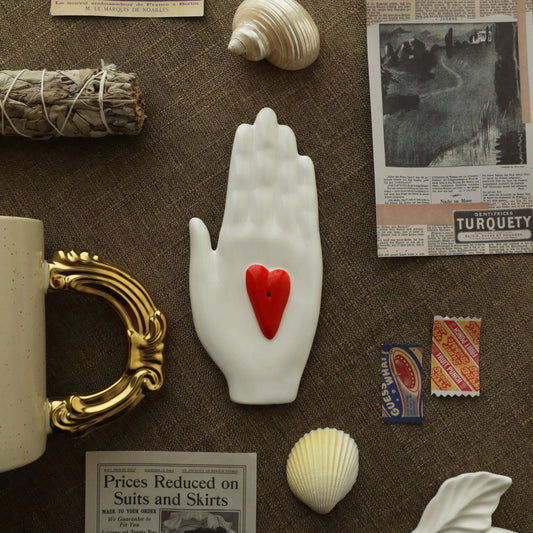 Overhead shot of a white ceramic hand-shaped incense holder with a red heart insert, arranged on linen with seashells, a smudge bundle, vintage stamps, and paper clippings.