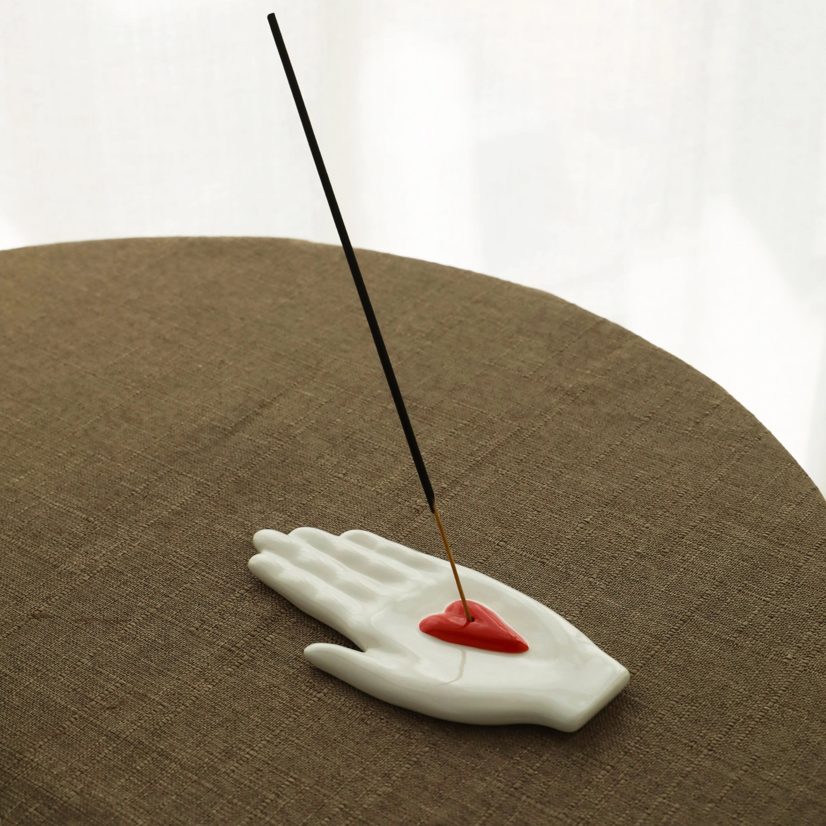White ceramic hand-shaped incense holder with a red heart insert holding a long incense stick on a linen-covered round surface, studio backdrop.