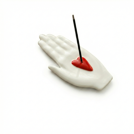 White ceramic incense holder shaped like an open hand with a red heart detail in the palm, holding a single black incense stick on a clean white background.