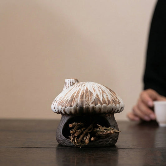Rustic ceramic mushroom house incense burner filled with dried wood twigs and natural herbs sitting on a wooden table with a hand holding a tea cup in the background