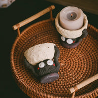 Handmade ceramic Soot Spirit candle holders displayed on a woven tray, with one holding a lit tealight candle.