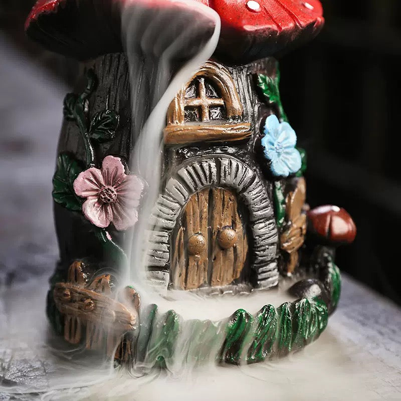 Close-up base details of the resin mushroom backflow incense burner, showing the mesmerizing thick smoke waterfall cascading down past a miniature fairy door, rustic stone textures, and vibrant pink and blue flower accents. Perfect for cottagecore room decor and aromatherapy.