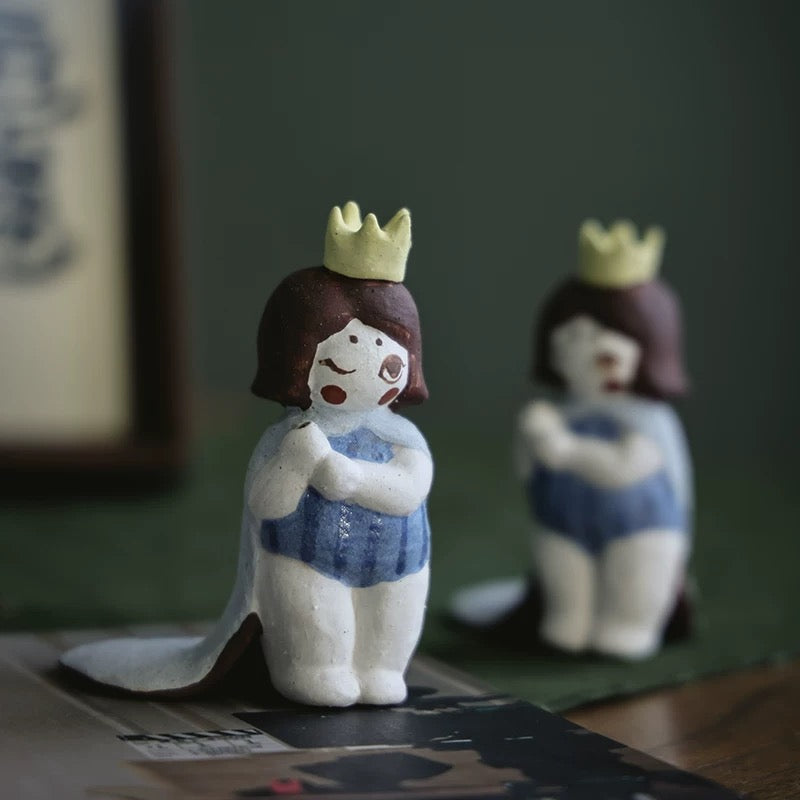 Handmade ceramic girl incense holder with yellow crown and blue outfit on stacked books; another figure blurred in the background, leaf-shaped tray behind.