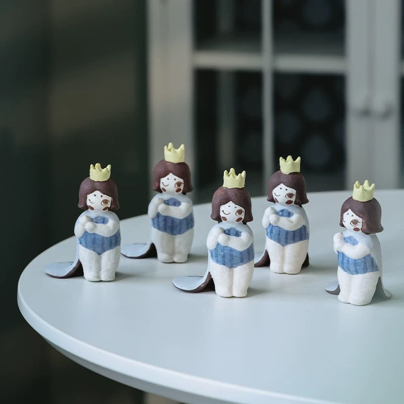 Group of five handmade ceramic girl incense holders with yellow crowns and blue outfits arranged on a round white table with leaf-shaped trays.