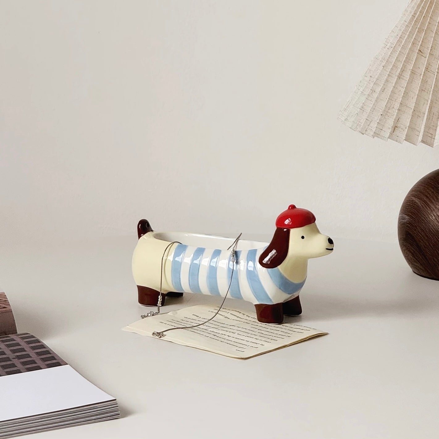 Ceramic dachshund incense holder in light blue stripes with a red beret, side view on a desk with papers and a lamp in the background.
