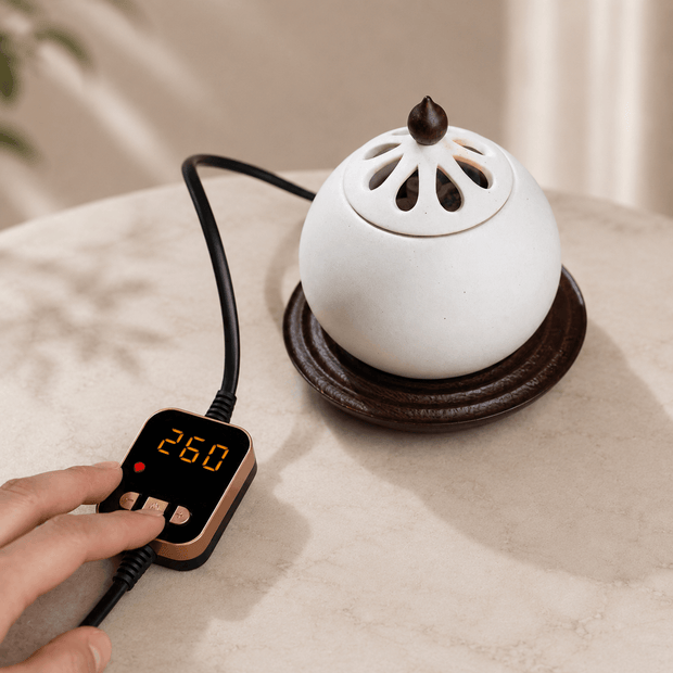A white ceramic incense burner with a perforated lid sits on a dark wooden base, connected to a digital temperature controller held by a hand. The scene is set on a light beige fabric surface with soft shadows.