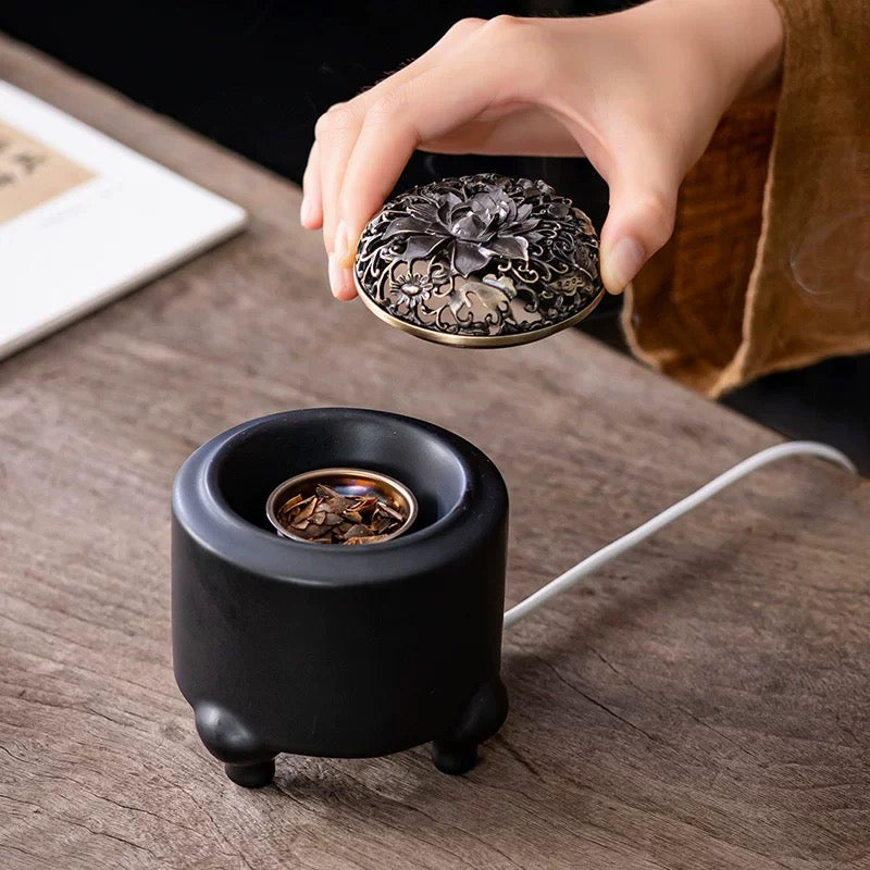 Electric incense burner with digital adjustable temperature and timer shown on a wooden tabletop, with a hand placing the ornate lid over the resin heating dish inside