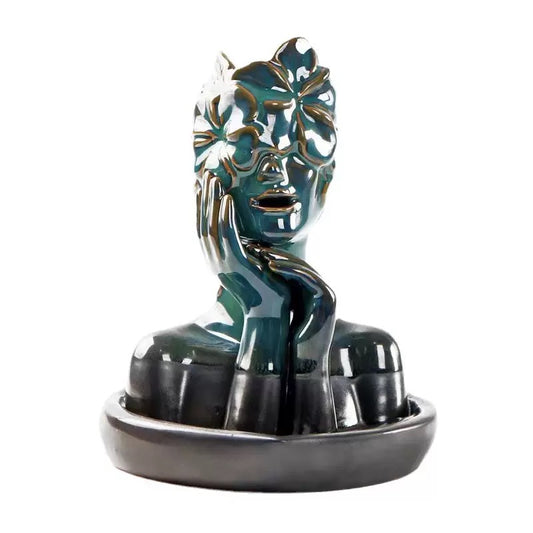 A glossy blue-green ceramic bust sculpture with raised floral mask details and a hand resting against the face, displayed on an oval base as an artistic incense holder and decorative accent.