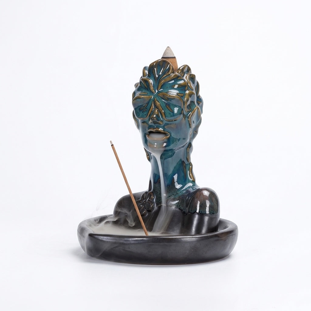 A glossy blue-green ceramic bust incense burner with floral mask details, featuring a backflow cone on top, white smoke flowing from the mouth into the oval base, and a stick incense holder at the side.