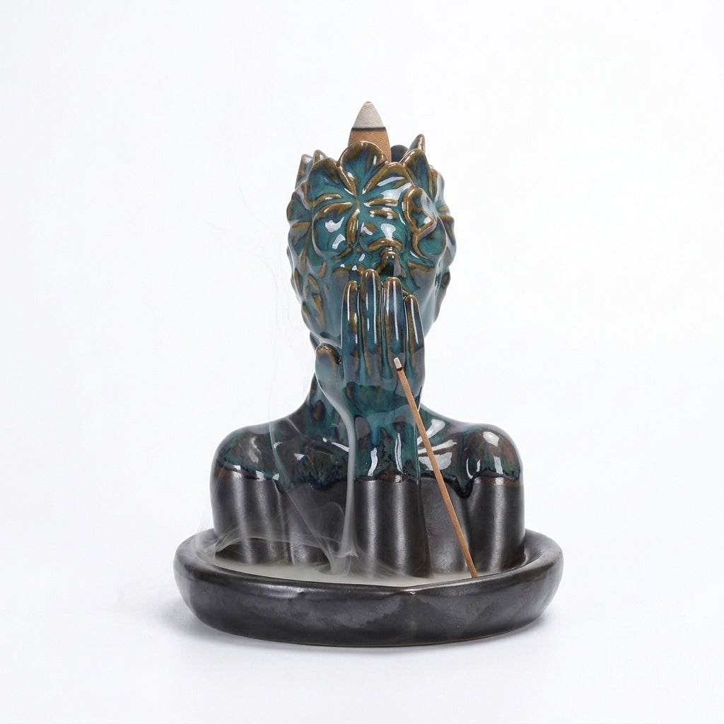 A glossy teal ceramic floral mask bust incense burner with a hand covering the mouth, a backflow cone on top, soft smoke drifting into the base tray, and a stick incense holder leaning at the front.