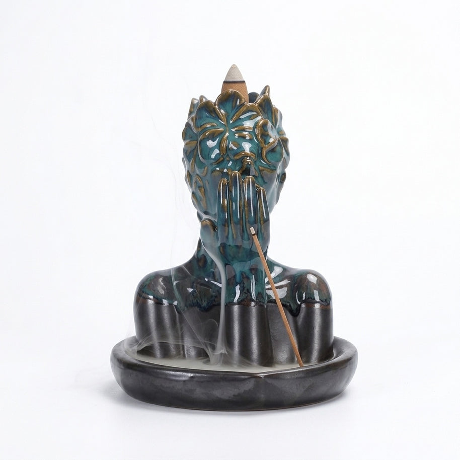 A glossy teal ceramic floral mask bust incense burner with a hand covering the mouth, a backflow cone on top, soft smoke drifting into the base tray, and a stick incense holder leaning at the front.